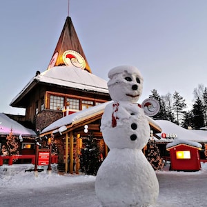 Könnte beinhalten: Ein großer Schneemann mit einem roten Schal steht vor dem Gebäude des Weihnachtsmannbüros in Finnland. Das Gebäude hat ein hohes, spitzes Dach mit einem kreisförmigen Logo, das den Weihnachtsmann und den Text Santa Claus Office Arctic Circle Finland zeigt.