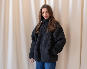 Può includere: Un maglione in pile grigio antracite scuro con collo alto e maniche lunghe, abbinato a jeans blu dal taglio ampio e scarpe slip-on in camoscio marrone. Il pile presenta un materiale testurizzato e soffice con chiusura a zip.