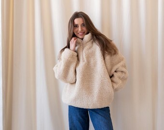 Può includere: Un maglione in pile beige a maniche lunghe, con collo alto e mezza zip frontale, abbinato a jeans blu dal taglio ampio e scarpe marroni senza lacci. Il maglione è realizzato in un materiale testurizzato simile allo sherpa.