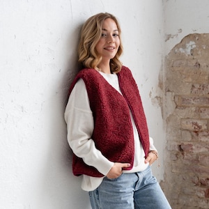 Può includere: Un gilet senza maniche color bordeaux in materiale pile testurizzato con scollo a V, indossato sopra una maglia bianca a maniche lunghe abbinata a jeans azzurri e scarpe da ginnastica bianche con accenti argento metallizzato.