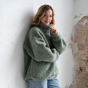 Peut inclure: Un pull en polaire vert sauge à manches longues avec un col montant et une fermeture éclair, associé à un jean bleu clair et des baskets de sport avec des accents blancs, argentés et beiges. La polaire présente une texture douce et duveteuse.