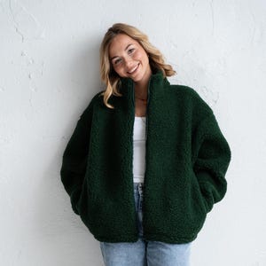 Puede incluir: Una chaqueta de forro polar sherpa en color verde bosque oscuro con cuello alto y cremallera frontal completa, usada sobre una camiseta sin mangas blanca lisa, combinada con pantalones vaqueros azul claro y zapatillas deportivas con detalles en blanco, gris y naranja.