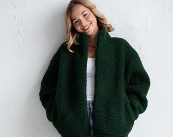 Può includere: Una giacca in pile sherpa verde bosco scuro con collo alto e cerniera frontale a tutta lunghezza, indossata sopra una canotta bianca semplice, abbinata a jeans in denim azzurro e scarpe da ginnastica con dettagli bianchi, grigi e arancioni.