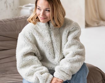 Può includere: Una giacca pullover a maniche lunghe e collo alto di colore grigio chiaro, realizzata in un materiale spesso, testurizzato e simile al pile. La giacca presenta una chiusura centrale con zip verticale che va dal colletto al centro del petto.