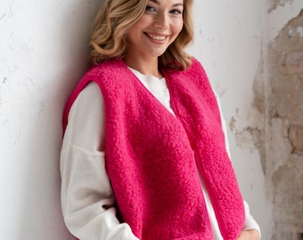 Può includere: Un gilet senza maniche rosa acceso realizzato in materiale pile testurizzato con scollo a V e chiusura frontale con zip, indossato sopra un maglione bianco a maniche lunghe e jeans azzurri.