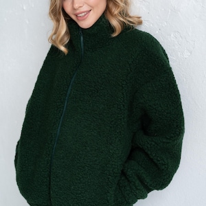 Puede incluir: Una chaqueta con cremallera de color verde bosque oscuro hecha de un material de forro polar sherpa grueso y texturizado. La chaqueta presenta un cuello alto, mangas largas y un cierre de cremallera frontal completo en un tono verde oscuro.