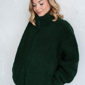 Puede incluir: Una chaqueta con cremallera de color verde bosque oscuro hecha de un material polar grueso y texturizado. La chaqueta presenta un cuello alto que sube por el cuello, mangas largas y un cierre de cremallera vertical central del mismo color.