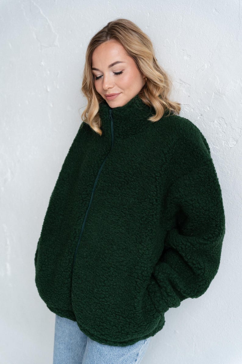 Puede incluir: Una chaqueta con cremallera de color verde bosque oscuro hecha de un material polar grueso y texturizado. La chaqueta presenta un cuello alto que sube por el cuello, mangas largas y un cierre de cremallera vertical central del mismo color.