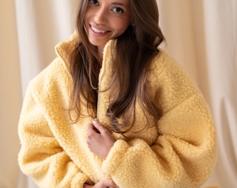Può includere: Una giacca con zip color giallo pallido realizzata in materiale pile testurizzato e soffice. La giacca presenta un colletto alto, maniche lunghe con polsini a coste e una chiusura frontale con zip integrale. È abbinata a jeans blu scuro.