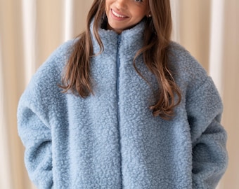 Puede incluir: Una chaqueta de forro polar de color azul claro, de manga larga, con cuello alto y cierre de cremallera vertical central. La prenda presenta un material grueso, texturizado y rizado con dos bolsillos laterales, sobre unos vaqueros azul oscuro.