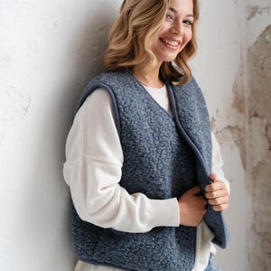 Può includere: Un gilet senza maniche realizzato in materiale pile riccio, blu e testurizzato. Il gilet presenta uno scollo a V ed è rifinito con un bordo blu liscio lungo i profili. È indossato sopra un maglione bianco a maniche lunghe e jeans azzurri.