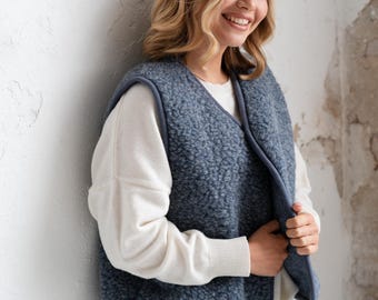 Può includere: Un gilet senza maniche realizzato in materiale pile riccio, blu e testurizzato. Il gilet presenta uno scollo a V ed è rifinito con un bordo blu liscio lungo i profili. È indossato sopra un maglione bianco a maniche lunghe e jeans azzurri.