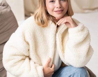 Puede incluir: Una chaqueta de color crema con cremallera y mangas largas, confeccionada en un material grueso, texturizado y similar al forro polar. La chaqueta presenta un cuello alto, una cremallera frontal completa y mangas holgadas, combinada con vaqueros azul claro.