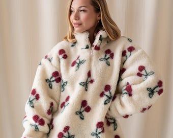 Può includere: Un maglione in pile color crema caratterizzato da un motivo ripetuto di ciliegie con frutti rossi e steli verdi. Il capo presenta un collo alto con chiusura a zip color argento, maniche lunghe con polsini a coste e una vestibilità ampia.
