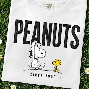 May include: A white t-shirt featuring a black graphic of Snoopy and Woodstock with the text PEANUTS in large black letters above them and the text SINCE 1950 below them. The shirt has a Comfort Colors brand label printed on the inside neck area.