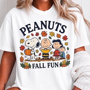 May include: A white short-sleeve t-shirt featuring a graphic of Snoopy, Charlie Brown, Lucy, and Linus surrounded by colorful autumn leaves and acorns. The text PEANUTS is printed in dark blue above the characters and FALL FUN below them.