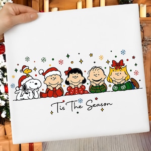 May include: A white square canvas featuring a colourful Peanuts holiday illustration. Snoopy, Woodstock, Charlie Brown, Lucy, Linus, and Sally are depicted lying down in a row. The text 'Tis The Season' is printed below them with small snowflakes.