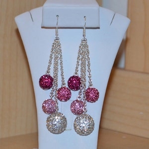 May include: Dangling earrings with silver chains and multiple crystal-covered spheres in shades of pink and silver. The earrings are displayed on a white jewelry stand, showcasing their cascading design and sparkling appearance.