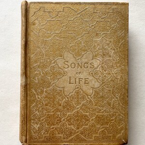 May include: A vintage book with a gold cover and intricate embossed designs. The title "Songs of Life" is printed in black lettering on the front cover.