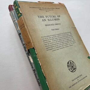 May include: A vintage book titled "The Future of an Illusion" by Sigmund Freud. The book is bound in green cloth with a gold spine and is part of the International Psycho-Analytical Library series. The book is in a third edition and has a price of 7s. 6d. on the spine.
