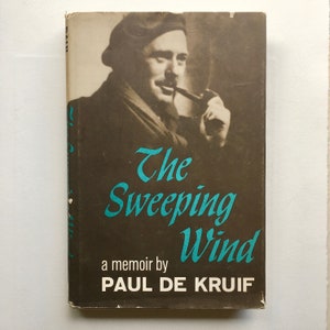 May include: A vintage book cover with a black and white photo of a man with a pipe. The title "The Sweeping Wind" is in teal blue lettering. The author is "PAUL DE KRUIF".
