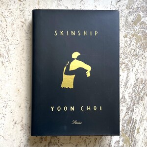 May include: A black book titled "Skinship" by Yoon Choi. The cover features a gold illustration of two figures embracing. The word "Stories" is printed in gold at the bottom of the book.