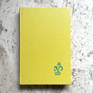 May include: A yellow hardcover book with a textured cover. The book has a teal emblem in the lower right corner with the letters "GHC". The book is closed and appears to be in good condition.