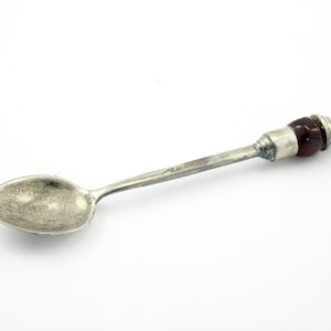 May include: A silver spoon with a dark red glass handle. The spoon has a rounded bowl and a long, slender handle.