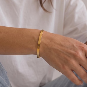 Può includere: Un bracciale rigido in metallo color oro lucido, sottile e con una superficie piatta e liscia. Il bracciale presenta un design semplice e minimalista che circonda il polso, mostrando una finitura metallica riflettente e una struttura uniforme.