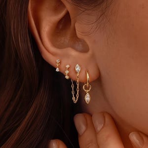 Pode incluir: Um conjunto de quatro brincos dourados usados numa única orelha. A coleção inclui um pequeno pino com pedra clara, um pino geométrico, um brinco de corrente com pedra lapidação marquise e uma argola dourada com pedra pendente.