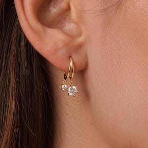 May include: A gold hoop earring featuring two dangling clear round-cut stones of different sizes. The earring is secured through the earlobe, showcasing a polished metallic finish and faceted gemstones that catch the light. The word Lexilinz is visible in the bottom right corner.