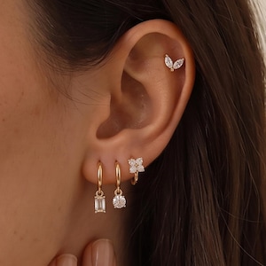 Könnte beinhalten: Ein Set goldfarbener Ohrringe mit einem blattförmigen Stecker mit zwei Marquise-Schliff-Steinen, zwei kleinen Creolen mit hängenden Baguette- und Rundschliff-Steinen sowie einem blumenförmigen Stecker mit vier runden Steinen. Der Text Lexilinz ist sichtbar.