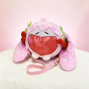 Puede incluir: Un bolso bandolera de felpa rosa claro, de forma redonda, con una ventana transparente en forma de corazón en la parte frontal. Tiene orejas largas y caídas decoradas con flores de cerezo bordadas y detalles de cerezas rojas colgantes.