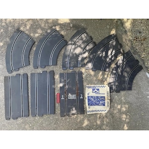 May include: A collection of black slot car track pieces, including curved and straight sections, along with a start/finish line piece and a small instruction sheet. The track pieces are made of plastic and have silver racing stripes.