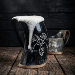 May include: A curved drinking horn mug with a dark, polished finish and a light-colored wooden base. The mug features a white engraved raven design and is filled with a frothy white liquid that spills over the rim. A second, smaller horn mug sits behind.