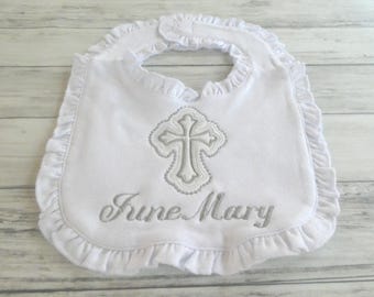 May include: A white baby bib featuring a ruffled edge and a hook-and-loop closure at the neck. The center is embroidered with a silver cross design and the name June Mary in a cursive script font, centered below the cross motif.