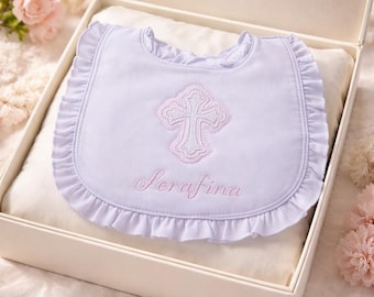May include: A white baby bib with a pink embroidered cross and the name "Serafina" in cursive. The bib has a ruffled edge and is presented in a white gift box with a soft, cream-colored interior.
