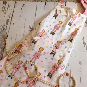 May include: A white apron featuring a repeating pattern of pink nutcracker figures and small colorful peppermint candies. The apron has pink neck ties and two front pockets. A string of natural wooden beads with colorful accents is draped over the fabric.