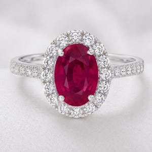 Puede incluir: Un anillo de tono plateado con un gran rubí rojo oscuro de corte ovalado en el centro. El rubí está rodeado por un halo de pequeñas piedras blancas redondas y transparentes. El aro también tiene una hilera de piedras blancas incrustadas.