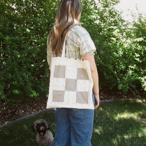 May include: A square tote bag featuring a patchwork design of cream-colored fabric and light brown checkered squares. The bag is bordered by a decorative scalloped eyelet lace trim and includes two matching fabric shoulder straps for carrying.