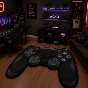 Puede incluir: Una alfombra de suelo grande y negra con forma de mando de videojuego. La alfombra presenta una cruceta direccional gris oscuro, dos joysticks circulares y cuatro botones de acción con símbolos de triángulo, círculo, cruz y cuadrado en color.