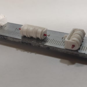 May include: Three small, white, cylindrical objects with red accents are arranged on a metal ruler. The objects appear to be components or parts, possibly for a larger mechanism or model. The ruler provides a scale for measurement.