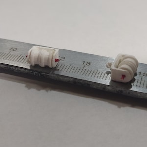 May include: Three small, white, coiled objects with red accents are positioned on a metal ruler. The ruler has centimeter markings. The objects appear to be components or parts, possibly for a larger device or model.