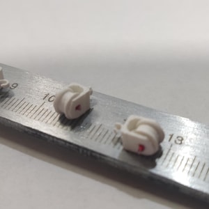 May include: Three small, white, plastic objects with a red dot are arranged on a metal ruler. The ruler has centimeter markings. The objects appear to be small mechanical components or decorative elements.