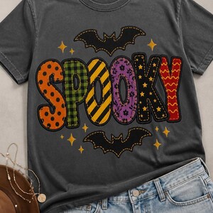 Op de afbeelding: Een antracietgrijs T-shirt met korte mouwen en het woord SPOOKY in grote, veelkleurige letters met patronen. Elke letter heeft een eigen ontwerp, waaronder stippen, ruiten, strepen en bloemen. Twee zwarte vleermuissilhouetten en gele sterren omringen de tekst.