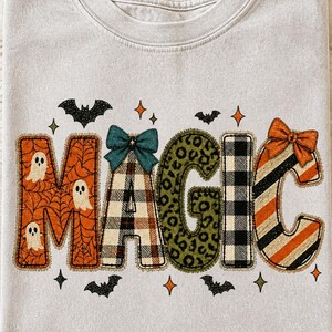 May include: A light gray t-shirt featuring the word MAGIC in large, patterned letters. The M is orange with spiderwebs and ghosts, the A has a teal bow and plaid pattern, the G is green leopard print, the I is black and white plaid, and the C is striped with an orange bow. Small black bats and orange stars surround the text.