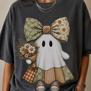 Puede incluir: Una camiseta gris carbón con el gráfico de un fantasma blanco que lleva un gran lazo floral con un botón en el centro. El fantasma sostiene un ramo de margaritas y flores naranjas, viste una falda de retazos y zapatillas de deporte naranjas.