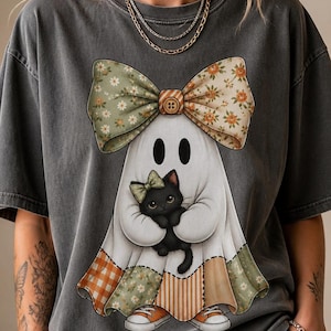 Könnte beinhalten: Ein anthrazitfarbenes T-Shirt mit einer Grafik eines weißen Geistes, der eine große Blumen-Schleife auf dem Kopf trägt. Der Geist hält ein kleines schwarzes Kätzchen mit einer grünen Schleife und trägt orange-weiße Sneaker. Der Geist hat einen Patchwork-Rock.