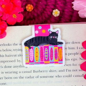 Puede incluir: Un marcapáginas de gato negro con una parte superior rosa de lunares y una base estampada colorida, apoyado en un libro. El marcapáginas presenta un gato sonriente y está diseñado para amantes de los libros.