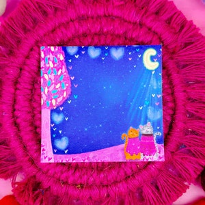 May include: A pink and blue illustrated card with two cats wearing sweaters and looking up at a crescent moon. The card is surrounded by a pink yarn wreath with white and pink pom-poms.
